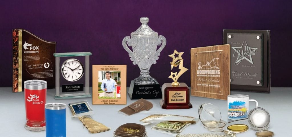 Various customized awards, plaques, trophies, and promotional items displayed on a table, showcasing high-quality engraving, signage, and personalized merchandise services.