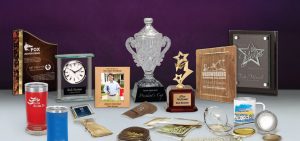Various customized awards, plaques, trophies, and promotional items displayed on a table, showcasing high-quality engraving, signage, and personalized merchandise services.