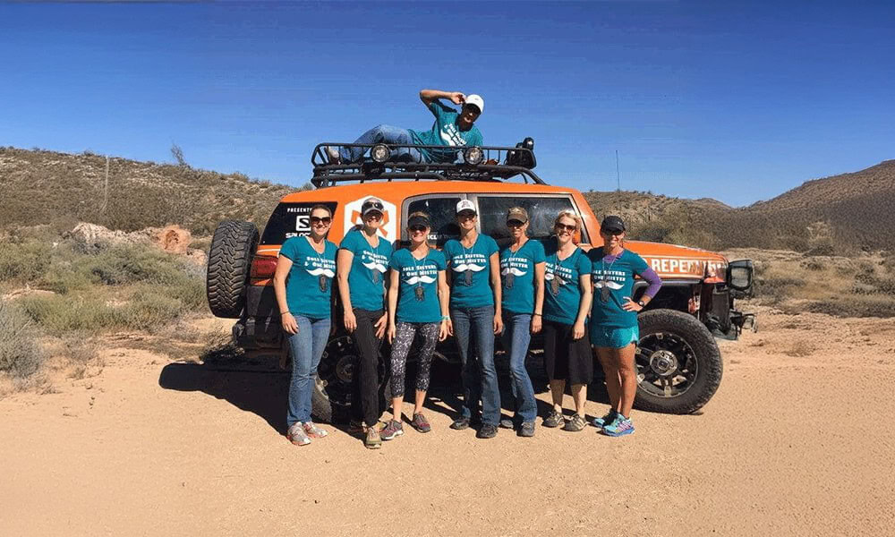 Group of women in matching teal T-shirts standing in desert front of off-road vehicle, team building outdoor activity, supportive colleagues, adventure, outdoor event, desert landscape, sunny day, rugged terrain.