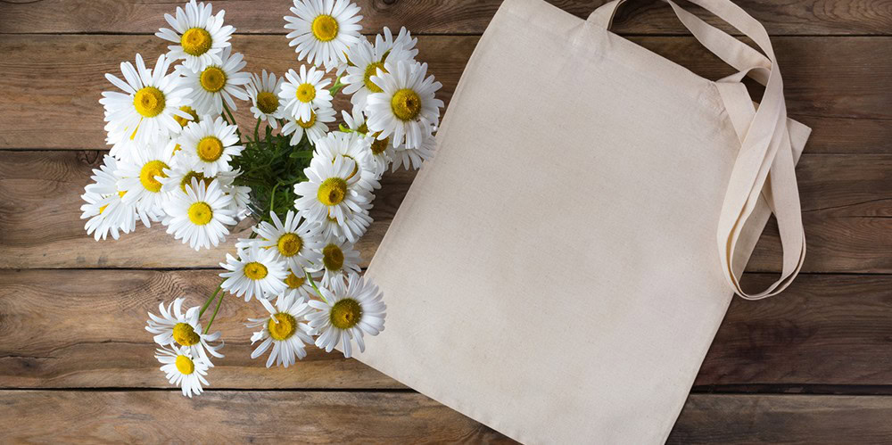 White daisies with yellow centers and a blank reusable tote bag on a wooden surface, ideal for custom printing or eco-friendly branding.