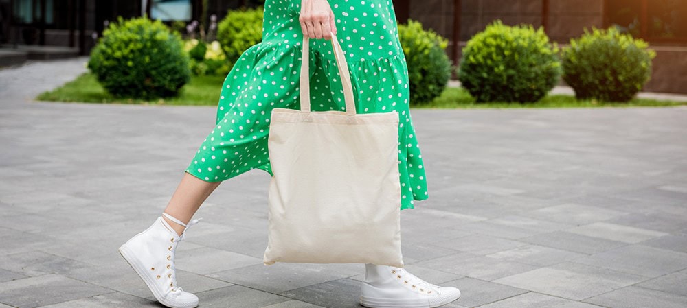 White canvas tote bag being carried by a person wearing a green polka dot dress and white sneakers, standing on an outdoor paved area with green bushes in the background.