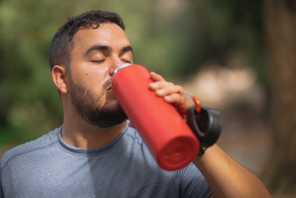 Man drinking water from red bottle outdoors, hydrating during exercise or hike.
