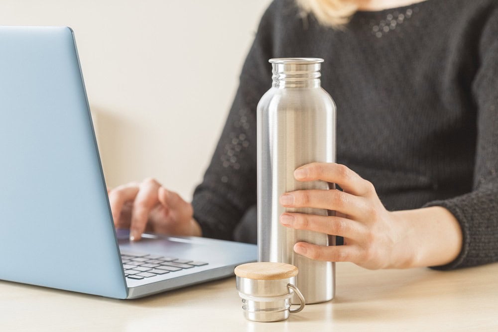 Stainless steel water bottle and laptop on a wooden desk, woman in black sweater working at home, eco-friendly beverage container, modern workspace accessories, health-conscious and environmentally friendly lifestyle.