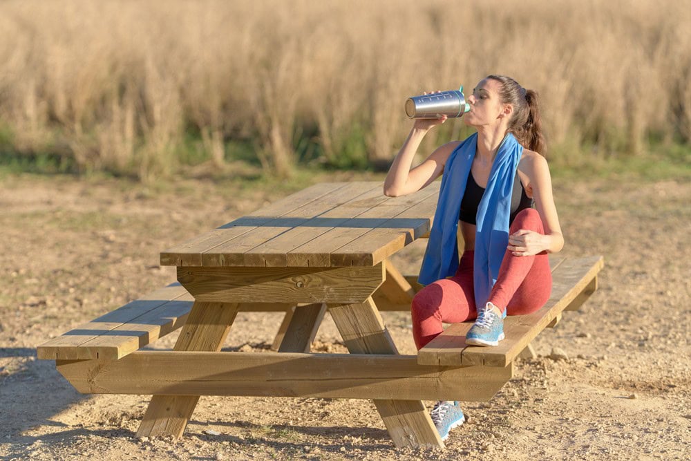 Refreshing outdoor beverage break on a sunny day, woman seated on a wooden picnic table drinking from a metal water bottle, dressed in athletic wear with a blue sweatshirt scarf, in a natural setting.