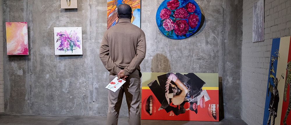 Why You Should (or Shouldn't) Print Your Photographs on Aluminum 1 Colorful art gallery featuring paintings and mixed media artwork displayed on concrete and brick walls, with a visitor observing and holding a brochure.
