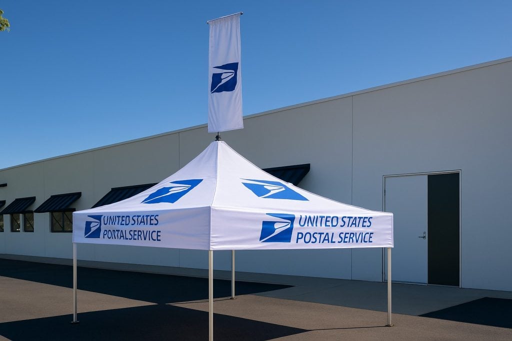 U.S. Postal Service branded canopy tent and flag set up outside a modern building with black window awnings, showcasing custom signs, banners, engraving, and apparel options for promotional and corporate use.