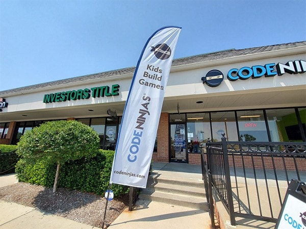Bright outdoor banner promoting kids build games for Codeninjas, displayed outside the storefront on a sunny day.