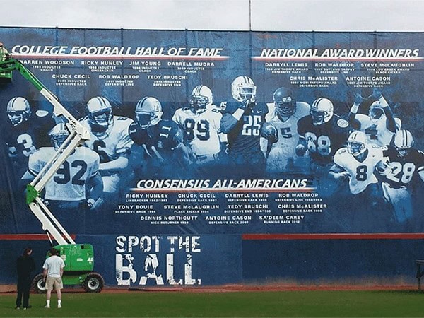 Large blue sports mural featuring football players, team names, and slogans, painted on an outdoor wall for a football team tribute or promotion. The scene includes a lift working on the mural.