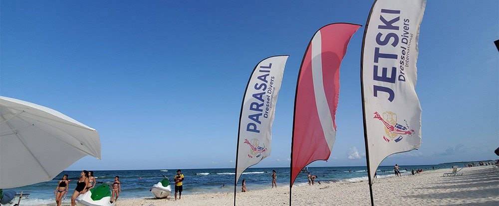 Bold, colorful beach flags with "Parasail" and "Jet Ski" branding flying on a sunny shoreline, showcasing marketing displays for water sports activities at the beach.