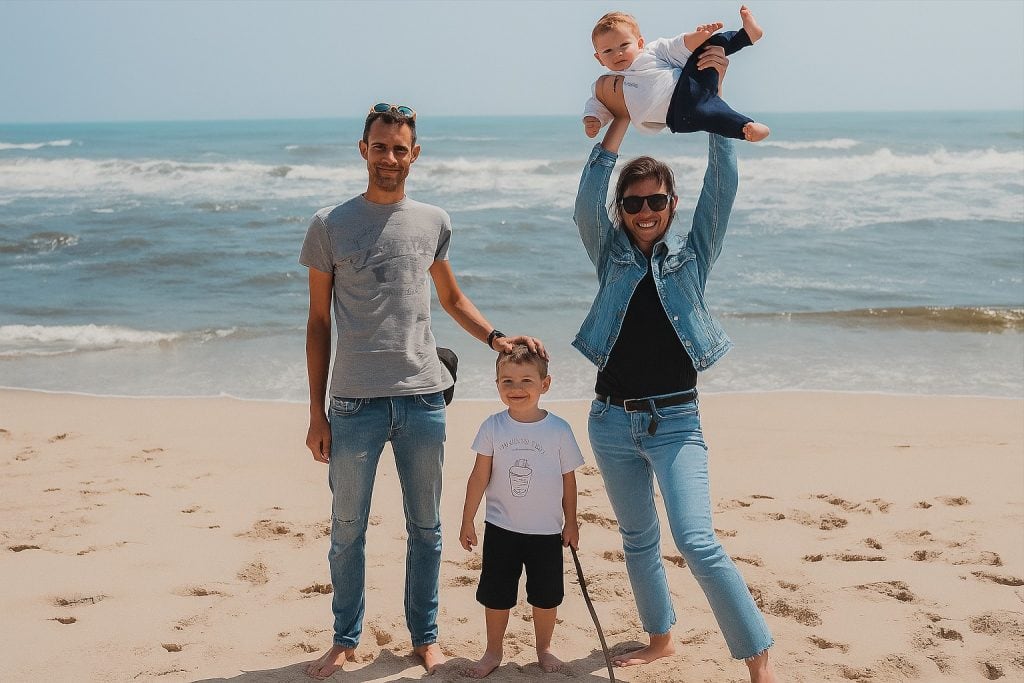 A happy family enjoying a day at the beach, with two adults, two children, and one child lifted in the air, all smiling and having fun on the sandy shore with the ocean in the background.
