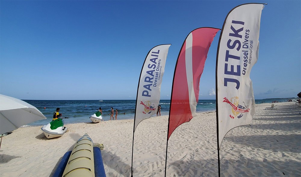 Feather Flag Design Tips - A Guide to Feather Flag Design 1 Colorful beach flags with jet ski and parasail advertising on a sunny sandy shore with people in the background, promoting water sports and beach activities.