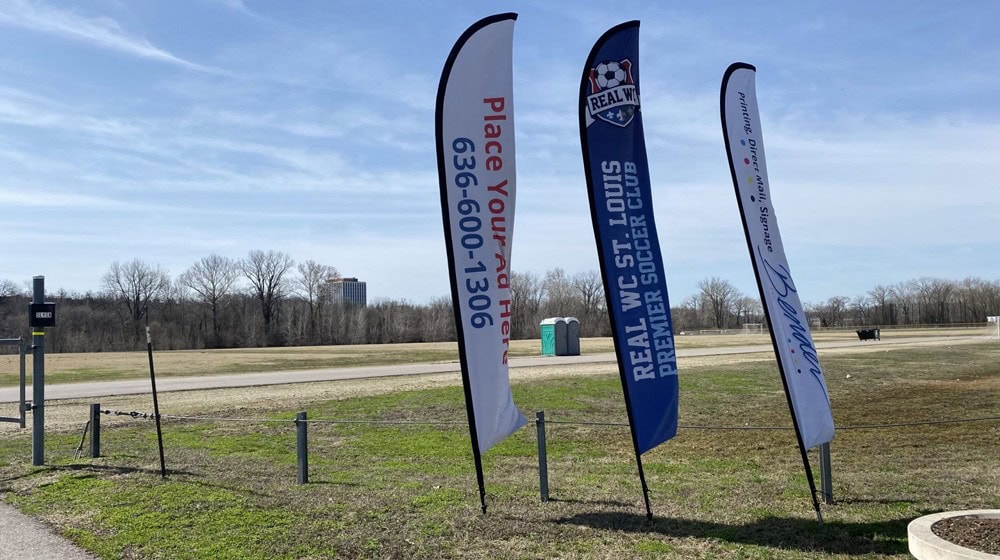 Feather Flag Design Tips - A Guide to Feather Flag Design 3 Vibrant advertising flags displayed outdoors along a grassy field with a clear blue sky, promoting local sports clubs and events with customizable banners and flags from Graphic Impact.