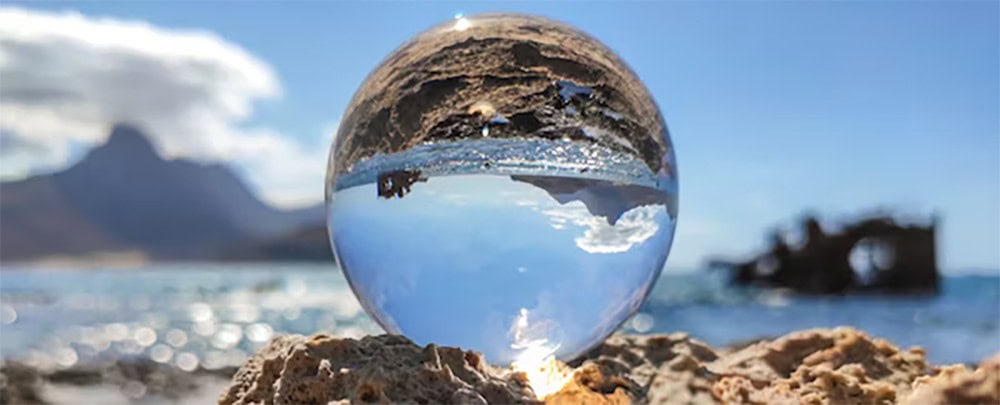 Glass sphere with mountain and ocean reflection on sandy beach in sunny weather.
