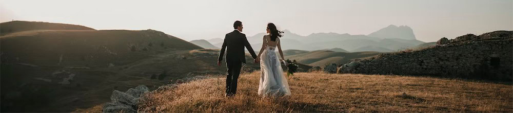 Elegant wedding couple walking hand in hand across scenic landscape at sunset, capturing romantic outdoor wedding moment.