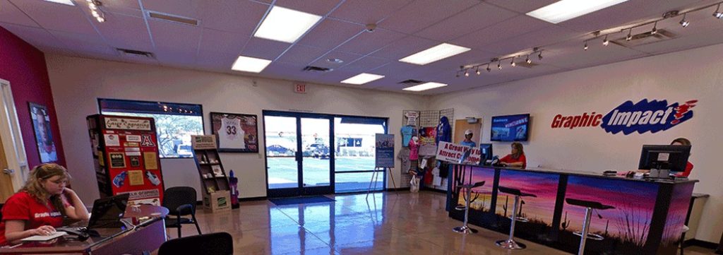 Bright interior of Graphic Impact signage and apparel shop showing a reception area with staff, display racks, and large windows; colorful signage, printed banners, and customer service desks.
