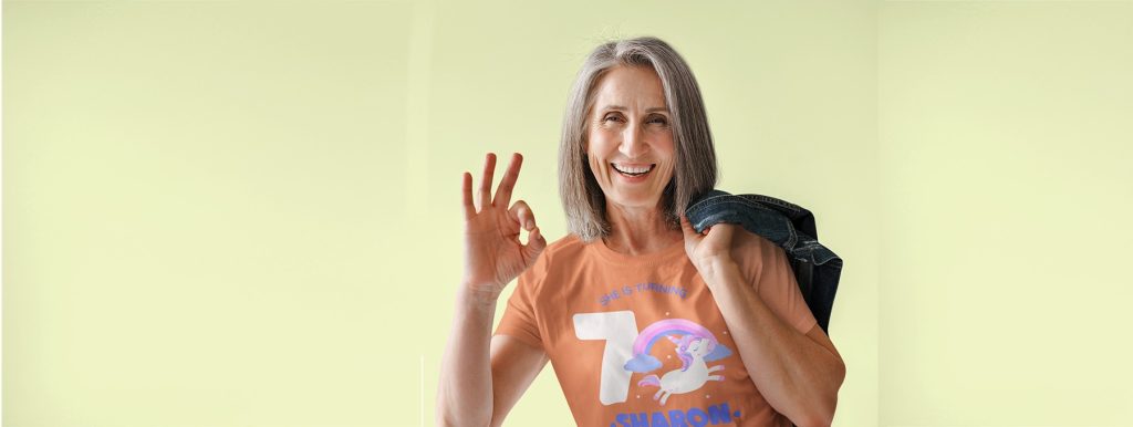 Celebrating a 70th birthday, smiling woman with gray hair, wearing a custom unicorn-themed shirt, holding a denim jacket over her shoulder, making an OK gesture, cheerful and vibrant atmosphere.