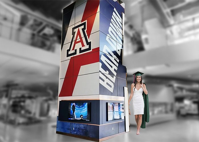 University of Arizona Wildcats logo banner with digital screens in an indoor space, featuring a woman in a graduation gown.