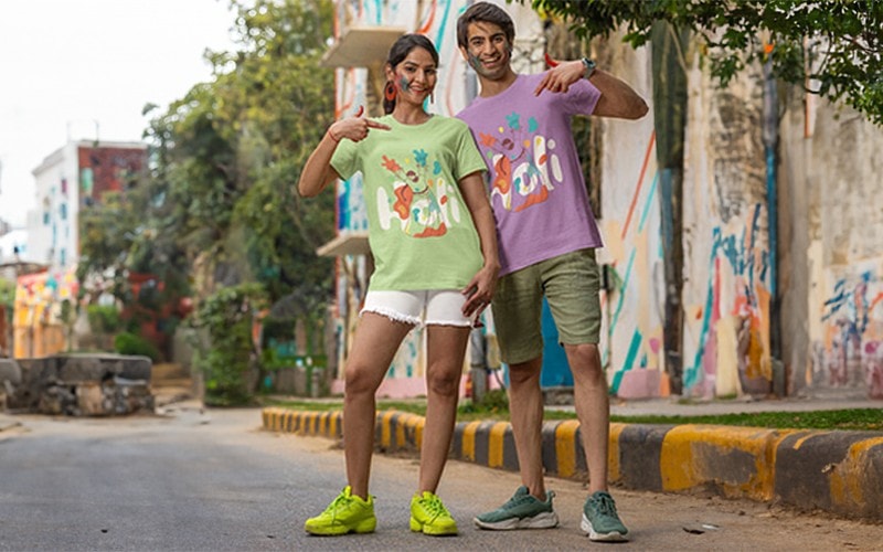 Colorful graphic t-shirts worn by smiling young adults standing outdoors in front of vibrant street art, showcasing custom apparel printing options at Graphic Impact.