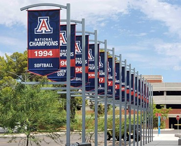 Colorful outdoor banners celebrating a softball team's national championship titles, displayed on flagpoles in a modern urban setting.