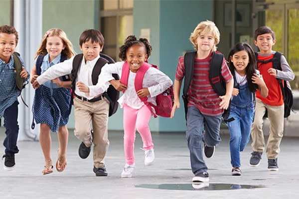 Brightly dressed elementary students running happily outdoors, carrying backpacks, in front of a school building.
